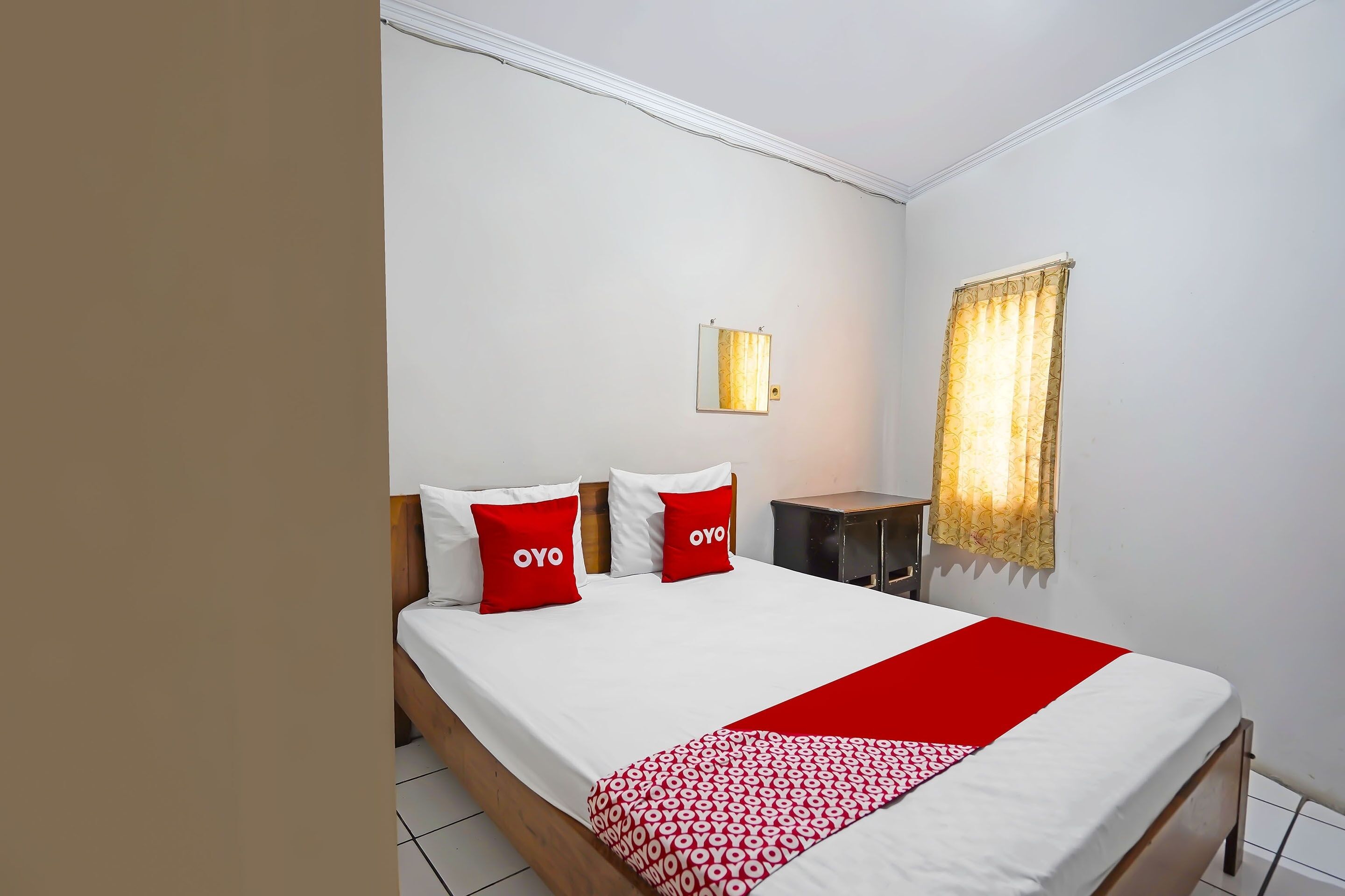 Une chambre d’hôtel avec un lit, deux oreillers rouges, une table de chevet, une lampe et une fenêtre avec des rideaux.