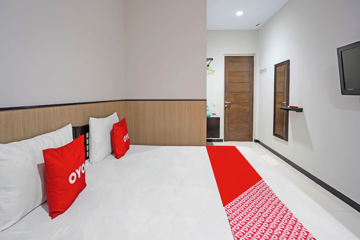 A hotel room with a bed, a wooden headboard, a red and white bed runner, a TV mounted on the wall, and a door with a window.
