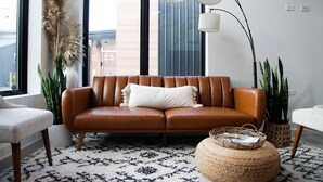 Ein modernes Wohnzimmer mit einem braunen Ledersofa, einem weißen Kissen, einem gemusterten Teppich, einem gewobenen Pouf und hohen Stehlampen.