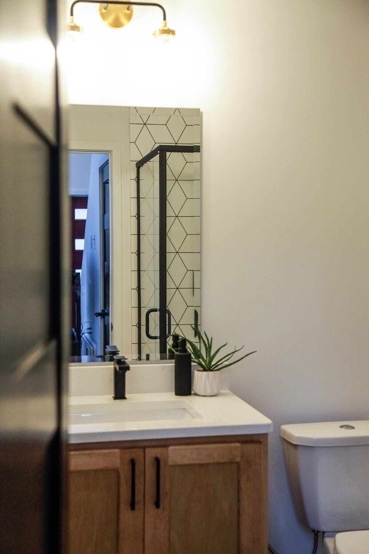 Une salle de bain avec un lavabo blanc, des meubles en bois, un miroir avec un motif géométrique en arrière-plan et des toilettes.