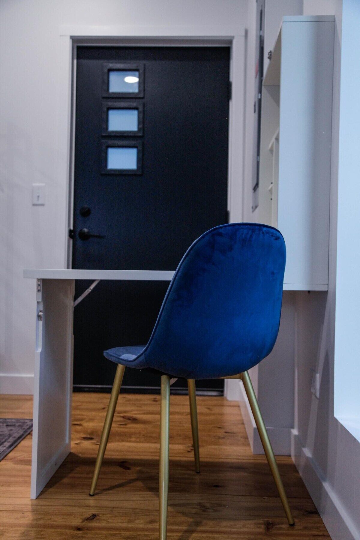 A blue chair with gold legs in a room with a wooden floor, a white desk, and a black door with a window.