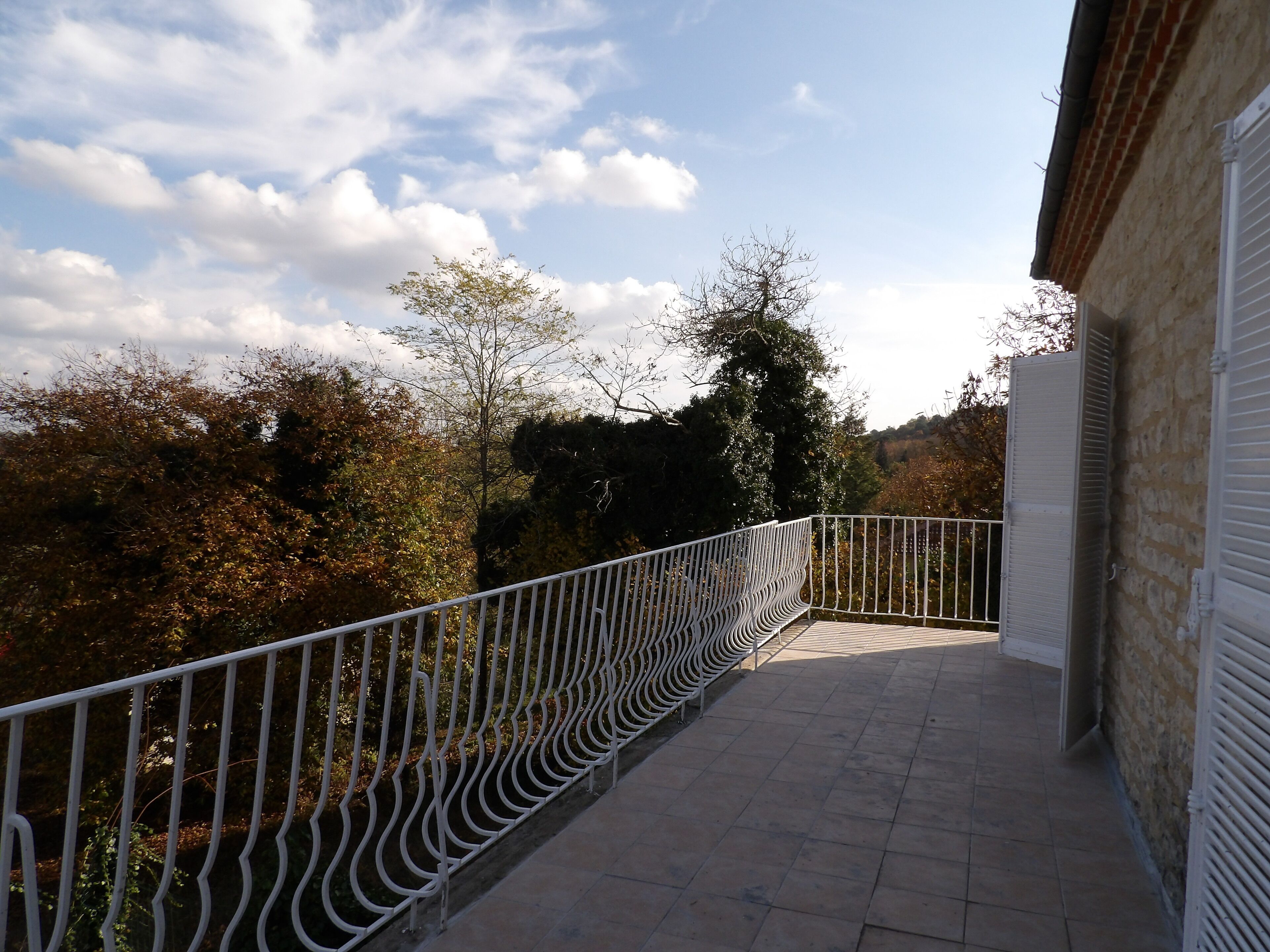 A balcony with a white metal railing, a tiled floor, and a view of trees and a clear sky.