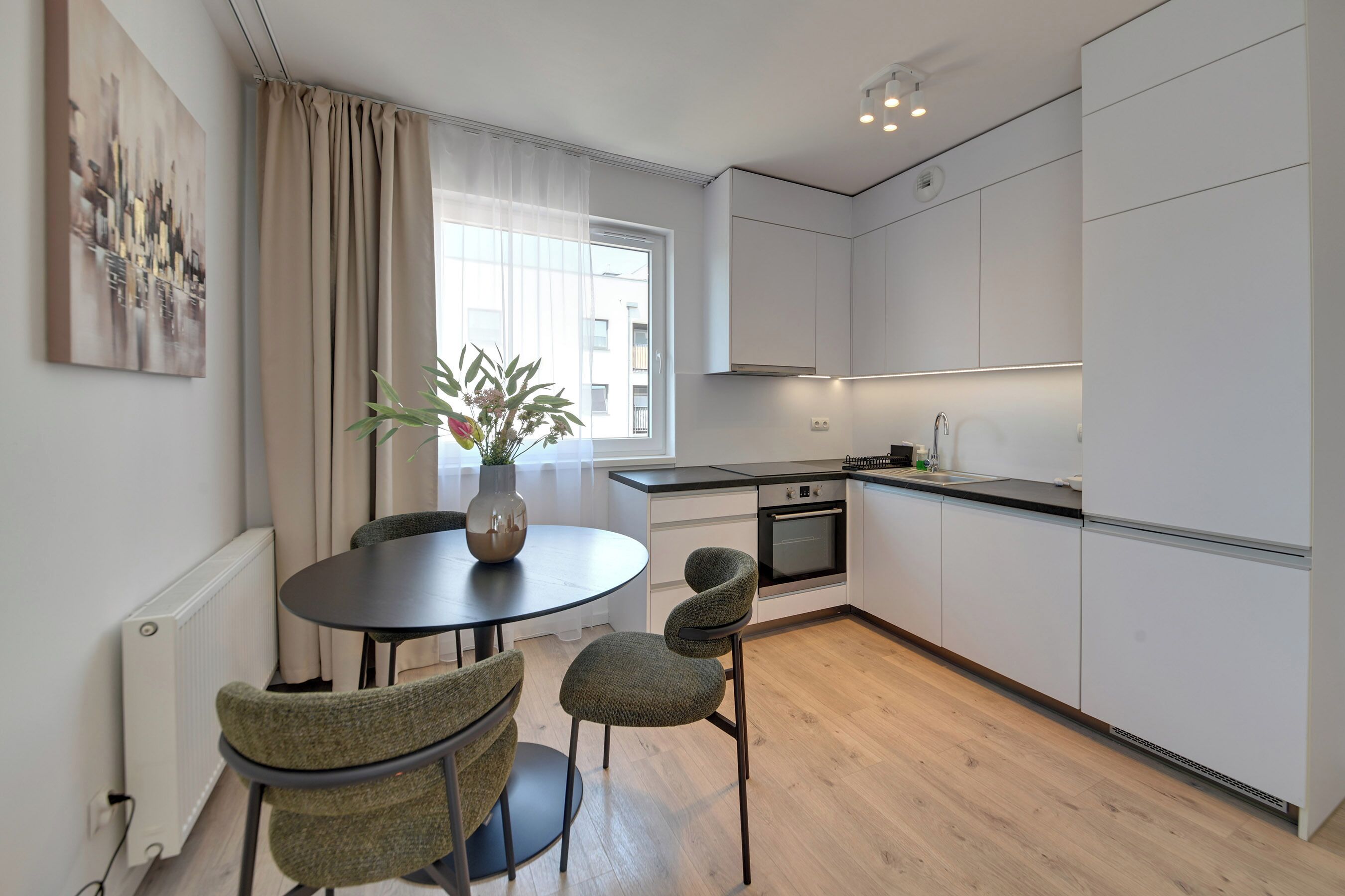 A modern kitchen with white cabinets, a dining table with chairs, a potted plant, and a window with curtains.