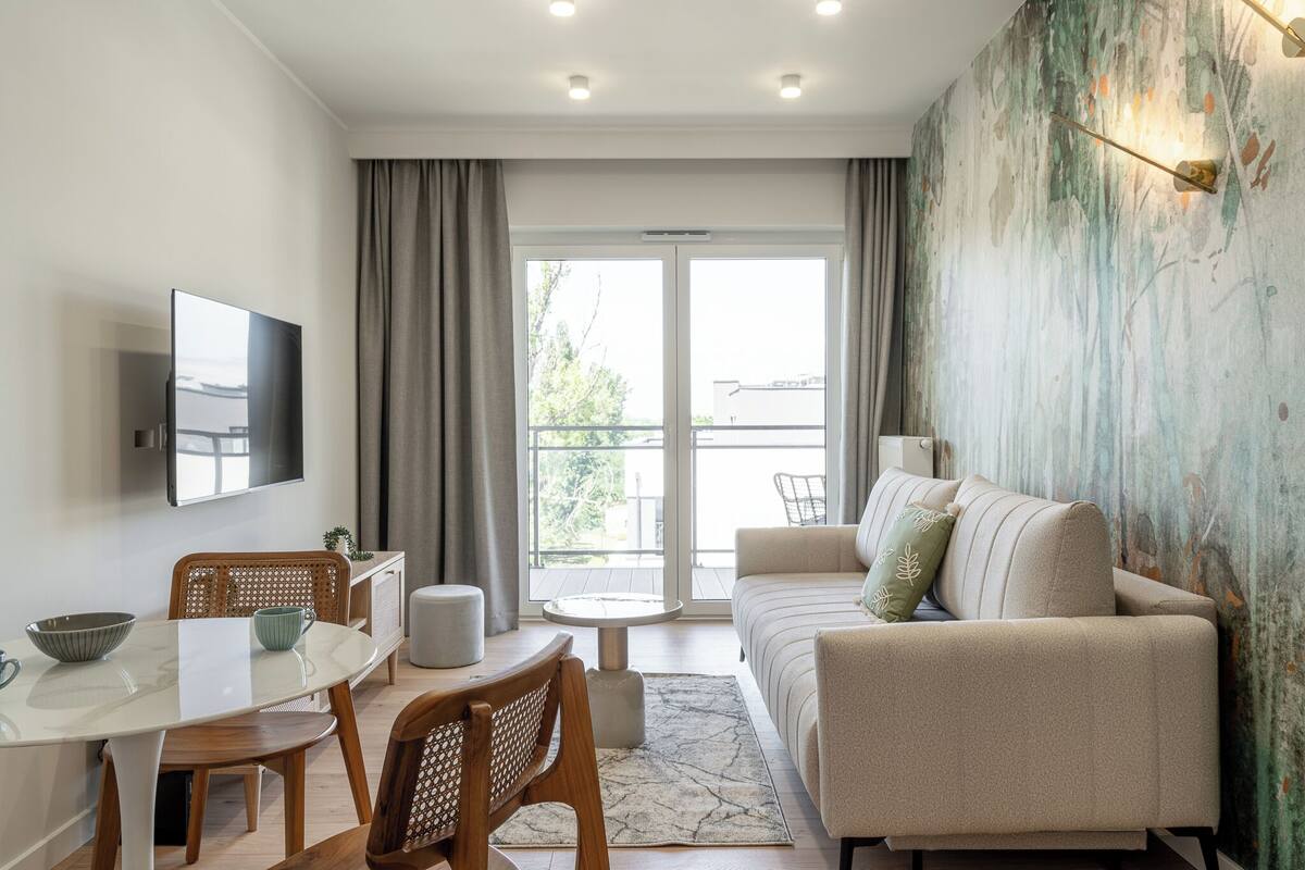 A modern living room with a sofa, dining table, and chairs. There is a TV mounted on the wall and a window with curtains.