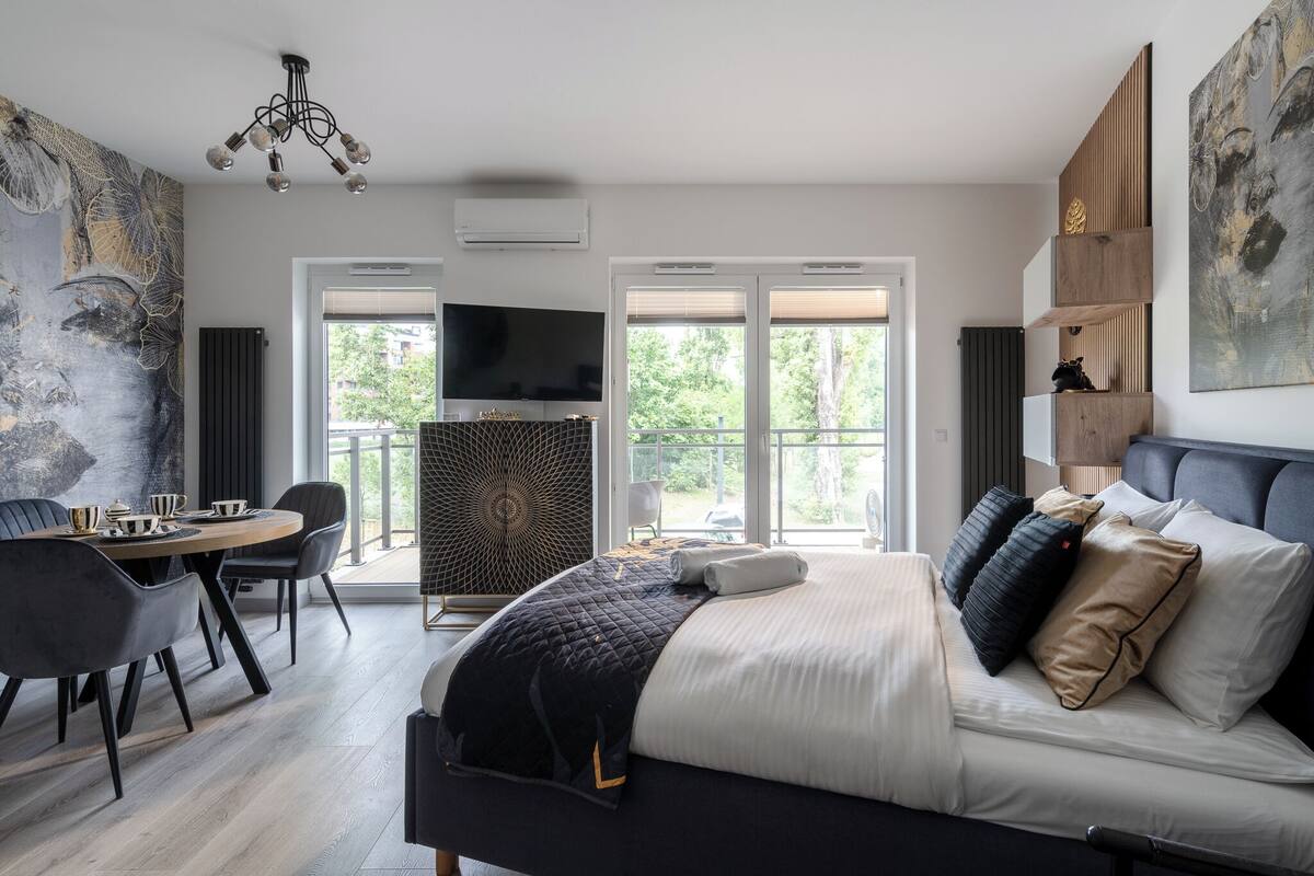 A modern hotel room with a large bed, a dining table with chairs, a TV, and a balcony with a view of trees.