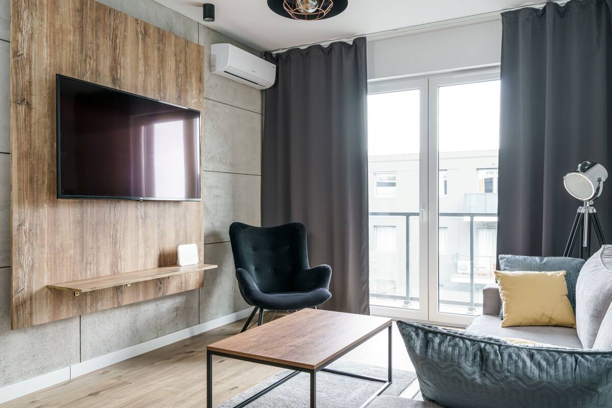 A modern living room with a flat-screen TV mounted on a wooden wall, a black armchair, a grey sofa with a yellow pillow, and a wooden coffee table.
