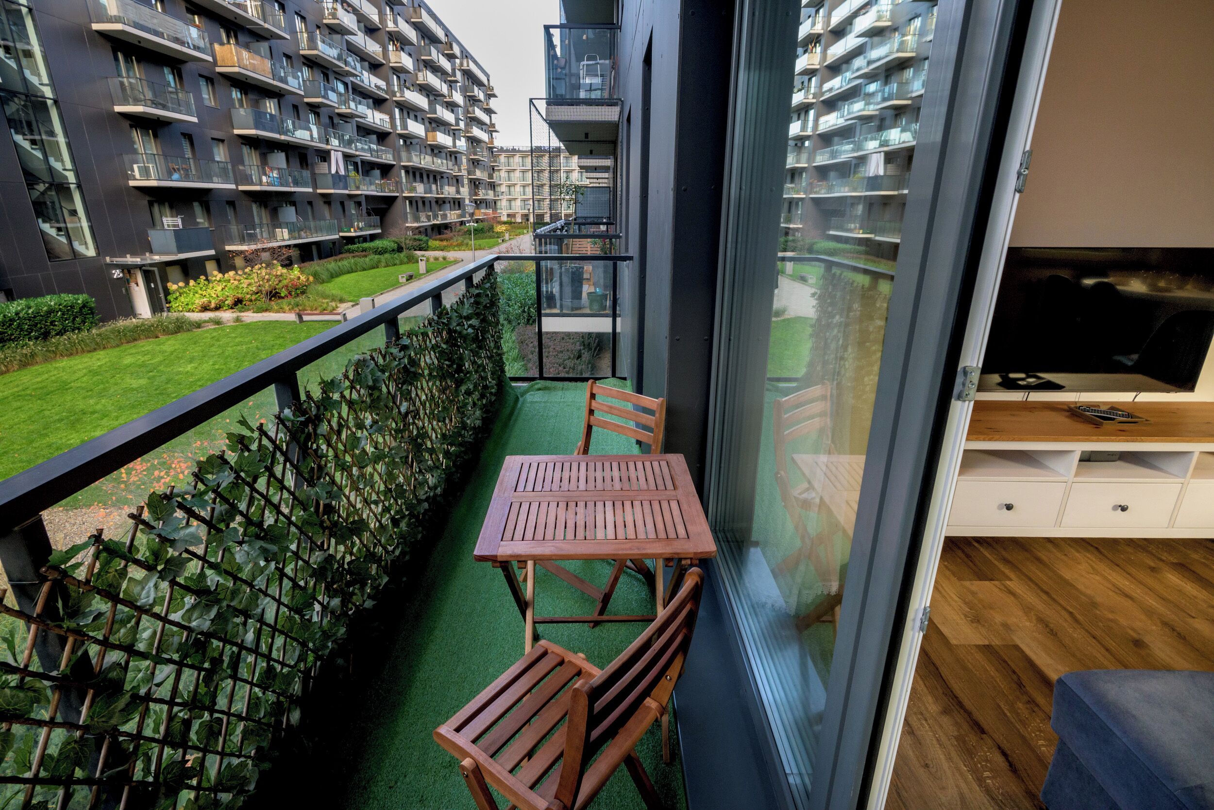 Un balcon avec une table et des chaises en bois, une vue sur un immeuble d’appartements moderne et un téléviseur posé sur un meuble en bois.