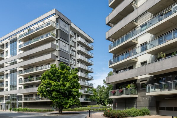 A modern multi-story residential building with balconies, large windows, and a tree in the foreground. - RentPlanet Kępa Mieszczańska (Wroclaw)