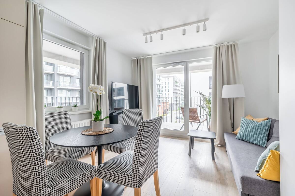 A modern living room with a round dining table, houndstooth patterned chairs, a sofa with cushions, a TV, and a balcony with a view of buildings.
