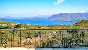 Un balcon doté d’une balustrade en métal, donnant sur un paysage côtier avec des maisons, une montagne et une étendue d’eau.