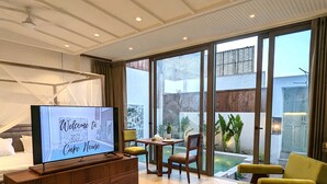 A modern living room with a television displaying "Welcome to Cape Nouse," a wooden entertainment unit, a dining area with a table and chairs, and large sliding glass doors leading to a garden. - Cape House at Amritsar (Amritsar)