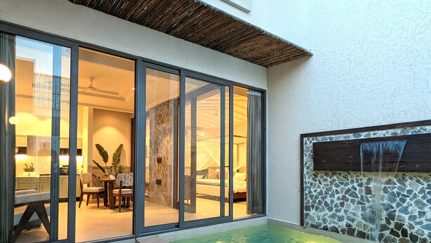 A modern hotel room with a glass door leading to a dining area, a small pool with a waterfall feature, and a stone wall. - Cape House at Amritsar (Amritsar)