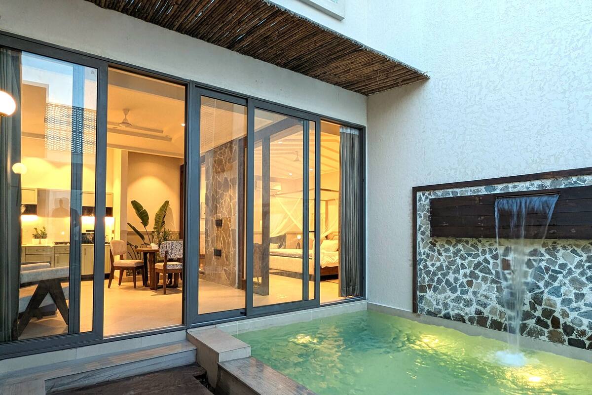 Una habitación de hotel moderna con una puerta de vidrio que conduce a un comedor, una piscina pequeña con cascada y una pared de piedra.