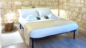 A bedroom with a stone wall, a bed with white linens, a bedside lamp, and a small wooden side table.