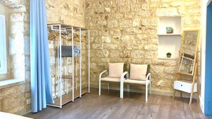 A room with stone walls, a wooden floor, a blue curtain, a metal shelving unit, a white chair with cushions, and a mirror on a stand.