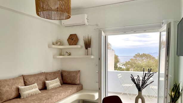 A modern living room with a beige sofa, a dining table with woven placemats, and a view of the sea through an open door. - Seaclusion (Santorini)