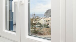 A window with white blinds, a view of a coastal area with buildings and a cliff. - Seaclusion (Santorini)
