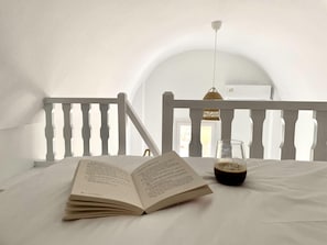 A book open on a bed with a glass of drink beside it, in a room with a white railing and a hanging light fixture. - Seaclusion (Santorini)