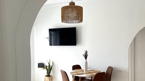 A dining area with a round table, chairs, a TV mounted on the wall, and a hanging light fixture. - Seaclusion (Santorini)