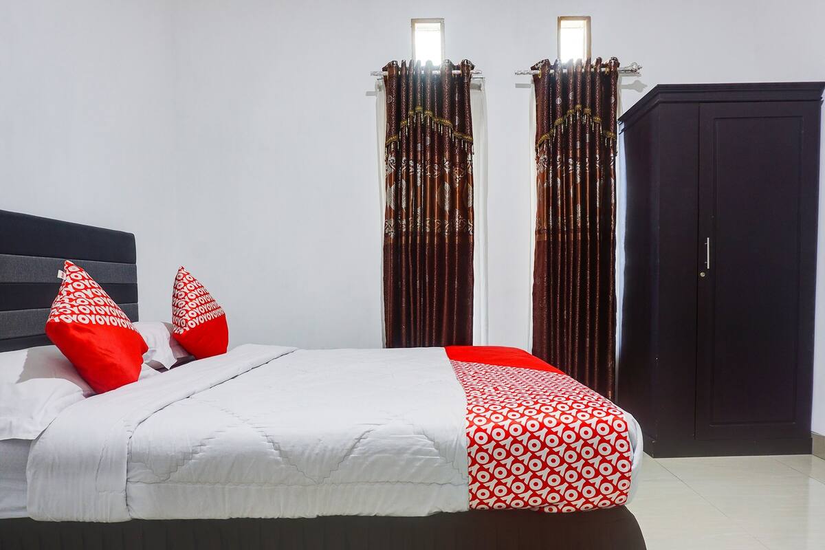 Una cama bien hecha con ropa de cama blanca y roja, dos almohadas rojas y un armario oscuro en una habitación con paredes blancas y una ventana.