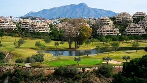 A golf course with a pond, surrounded by residential buildings and mountains in the background. - Los Piños, 2 Bedroom Apartment with panoramic view (Benahavis)