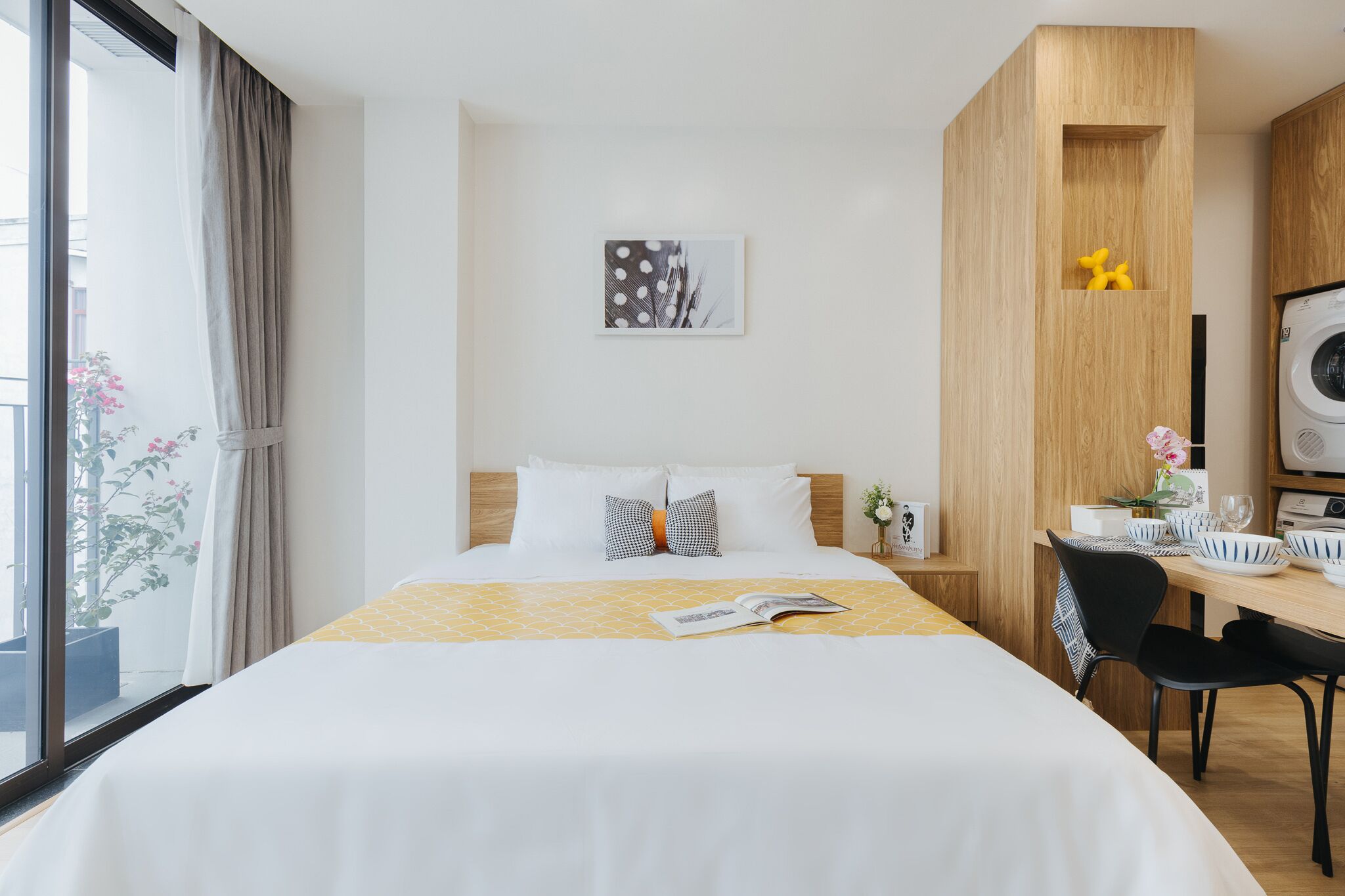 A hotel room with a bed, a desk with a chair, a window with curtains, and a wall-mounted artwork.