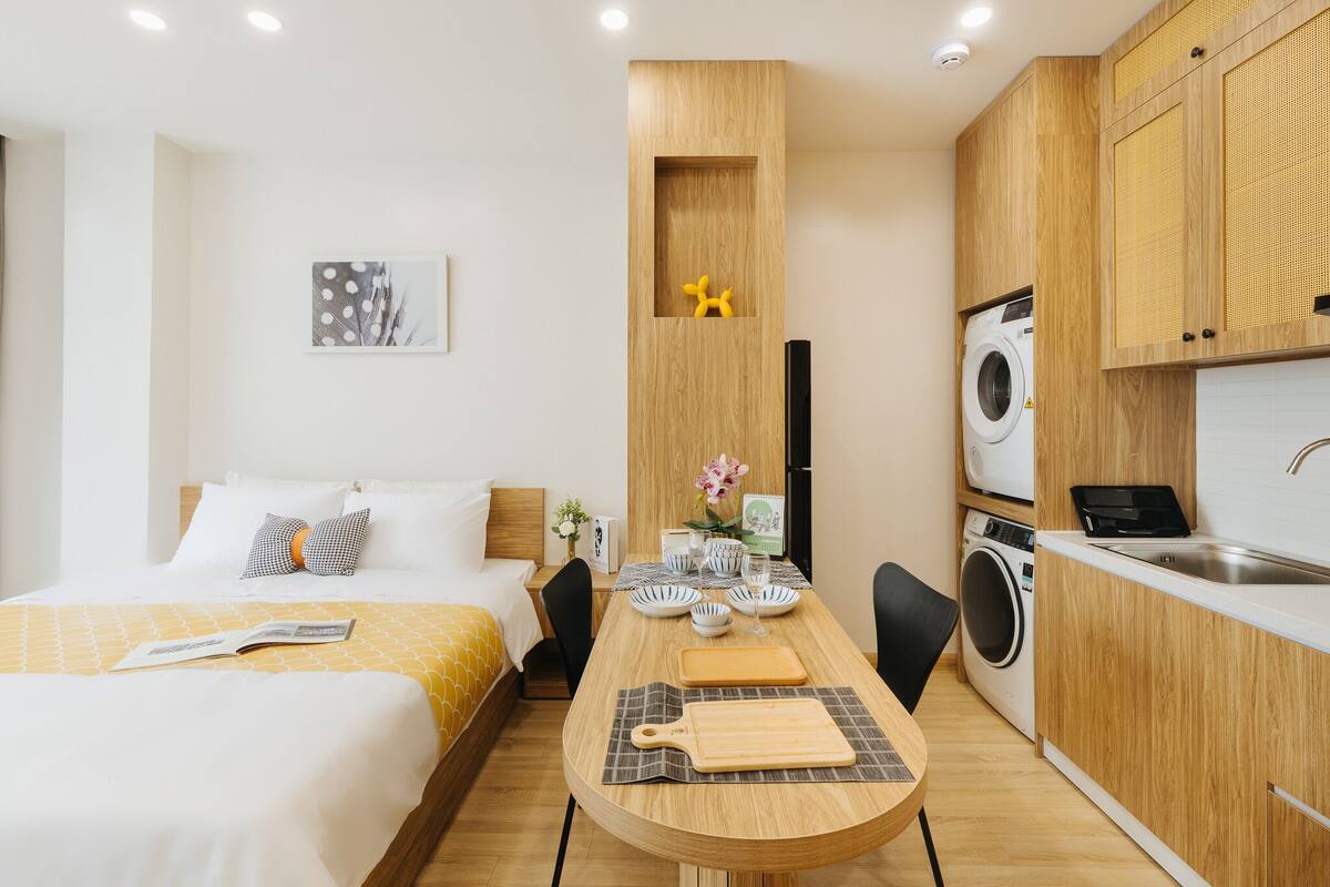 Quarto de hotel moderno com cama, mesa de cabeceira e uma pequena cozinha compacta, incluindo uma máquina de lavar roupa.