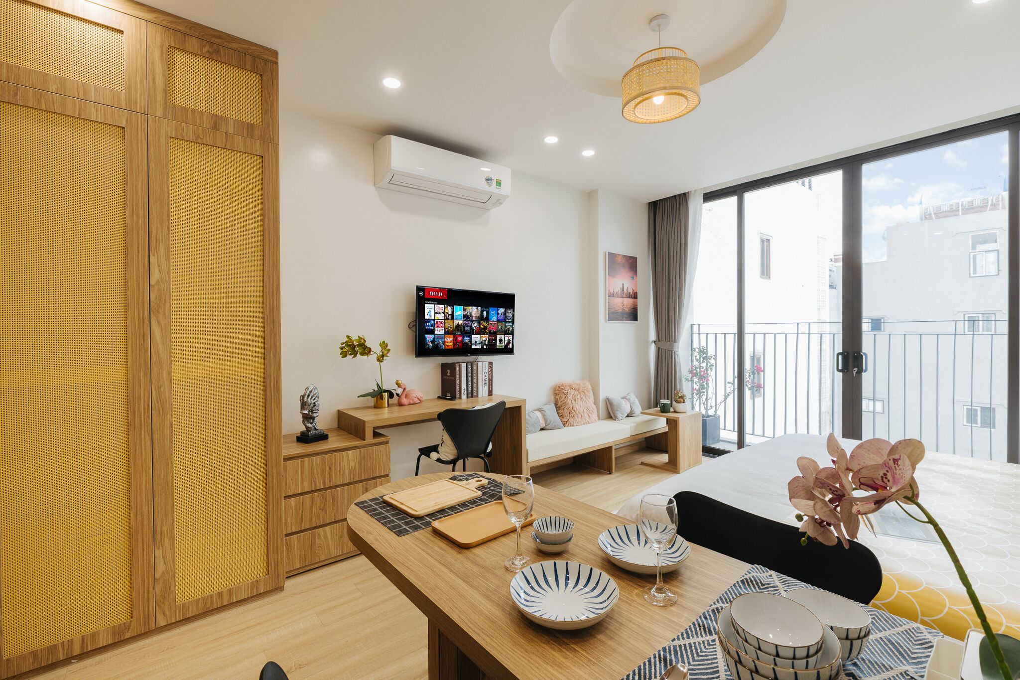 A modern hotel room with a wooden wardrobe, a dining table with plates and glasses, a TV on the wall, and a balcony with a view.