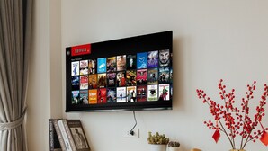 A modern living room with a flat-screen TV displaying a Netflix interface, a wooden desk with a chair, a small potted plant, a decorative vase with red berries, and a hat. - Myrcella Urban Studio (Hanoi)