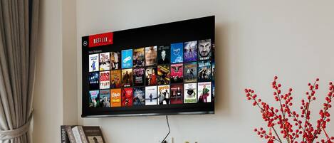 Moderni olohuone, jossa on seinälle asennettu taulutelevisio, jossa näkyy Netflixin käyttöliittymä, puinen työpöytä tuolilla, pieni ruukkukasvi, koristeellinen maljakko, jossa on punaisia marjoja, ja hattu.