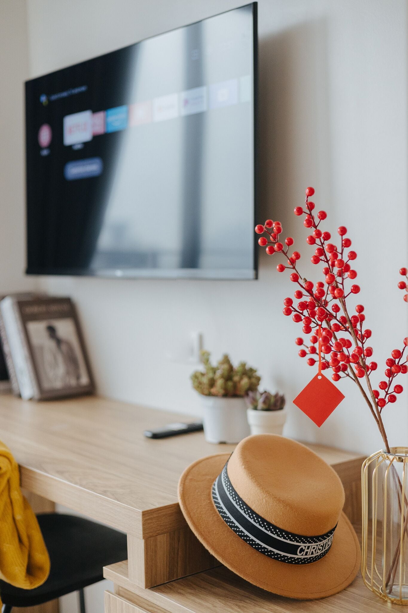 Una sala de estar moderna con un escritorio de madera, un televisor montado en la pared, un cuadro enmarcado, una pequeña planta en maceta y una rama decorativa con bayas rojas.