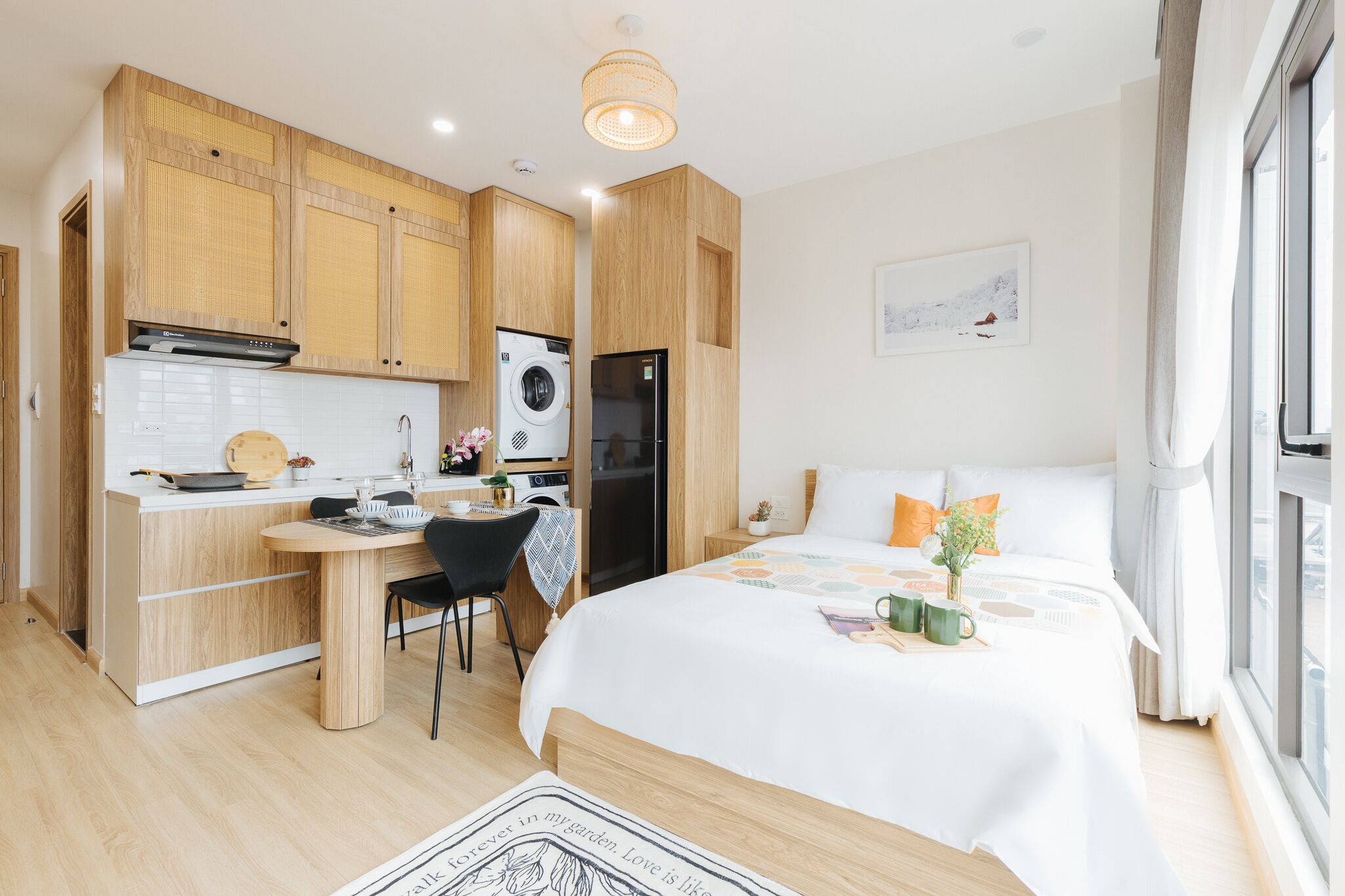 A modern, compact hotel room with a kitchenette, a bed with a white duvet, and a large window providing natural light.