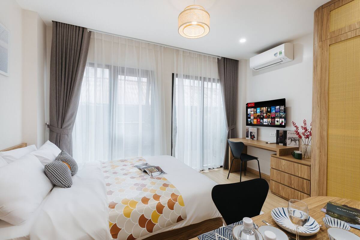 Quarto de hotel com cama, escrivaninha, cadeira, televisão e área de refeições com pratos e um copo.