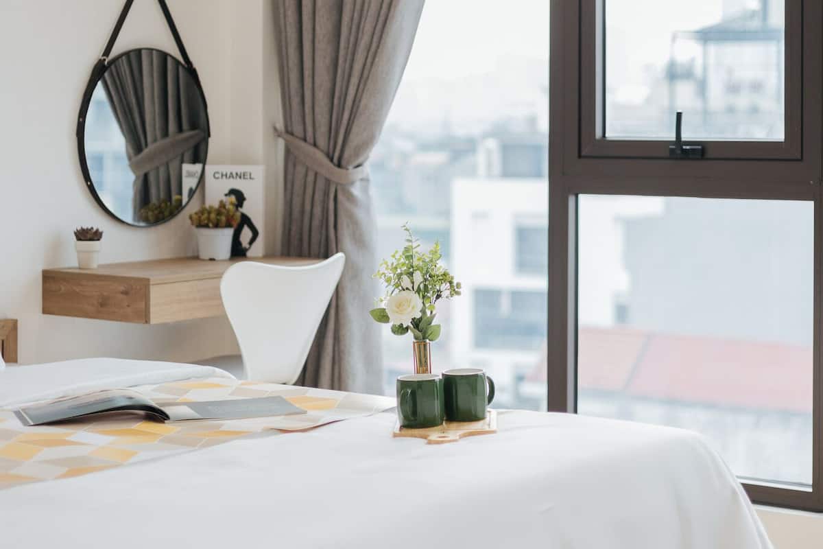 Quarto com uma janela grande, uma cama, mesa de cabeceira com espelho, uma planta pequena e uma caneca verde.