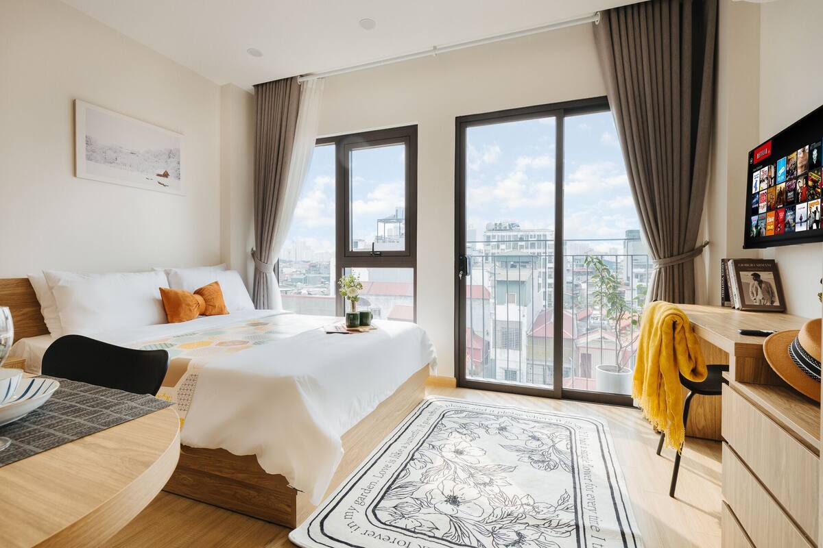 Quarto de hotel moderno com uma cama grande, uma escrivaninha, uma cadeira e vista para a cidade através de amplas janelas.