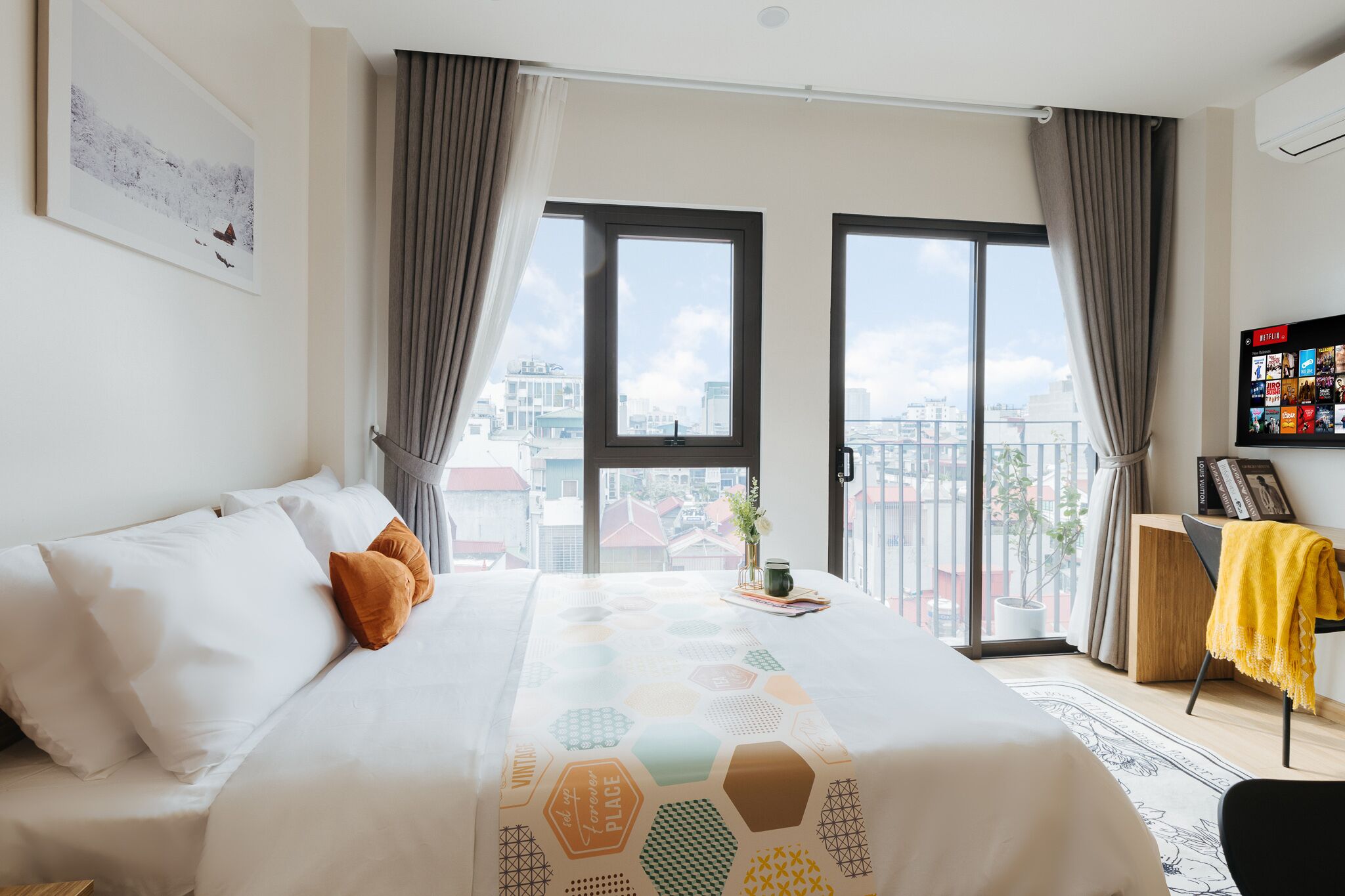 Camera d'albergo moderna con un letto grande, vista sulla città attraverso ampie finestre e un piccolo tavolino con una pianta.