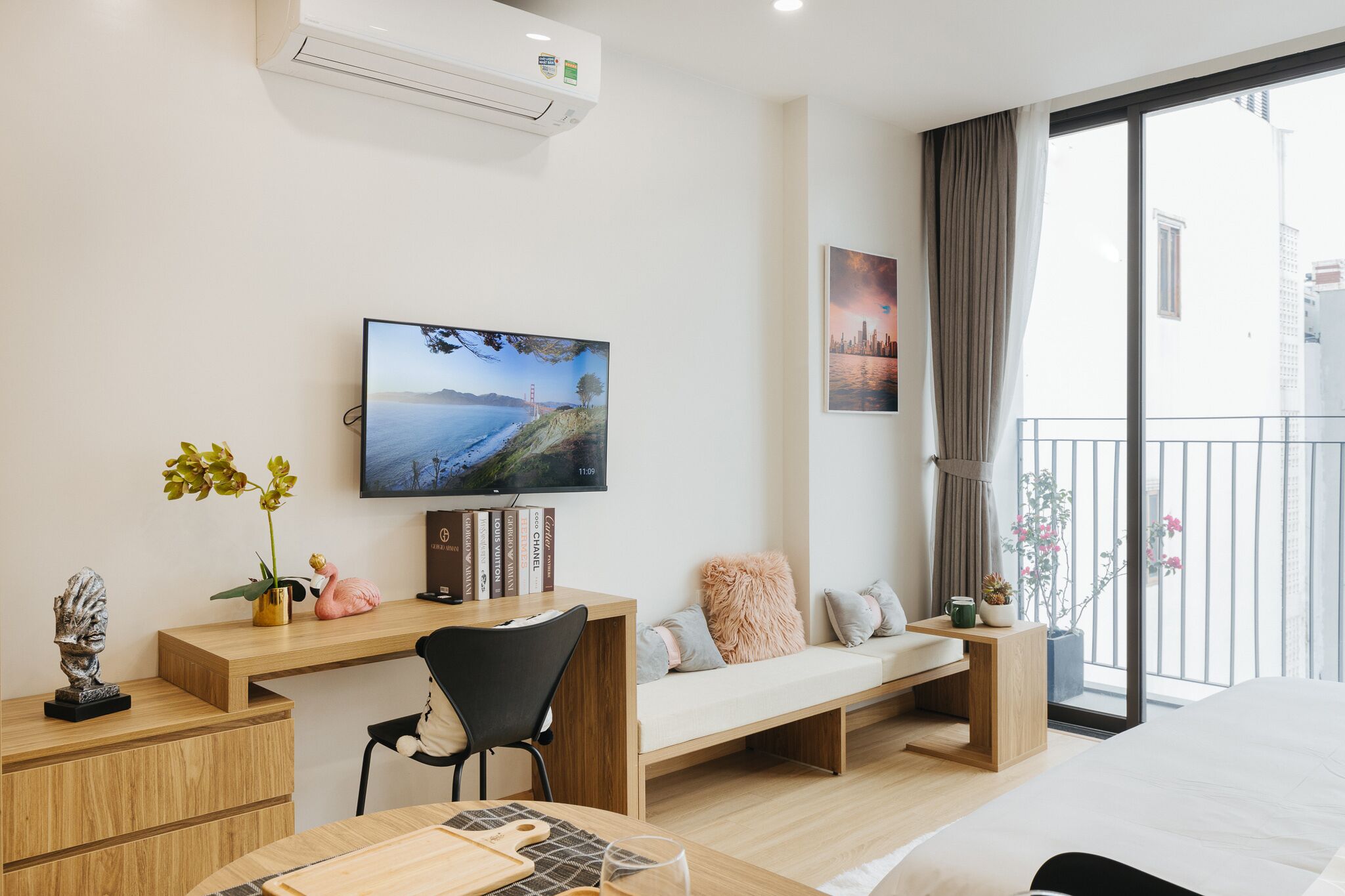 A modern hotel room with a wooden desk, a comfortable sofa, a bed, a TV mounted on the wall, and a balcony with a view of the city.