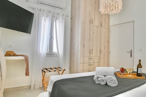Habitación de hotel con cama, televisor montado en la pared, armario de madera, una mesita con bandeja de toallas y vasos, y una botella de vino.