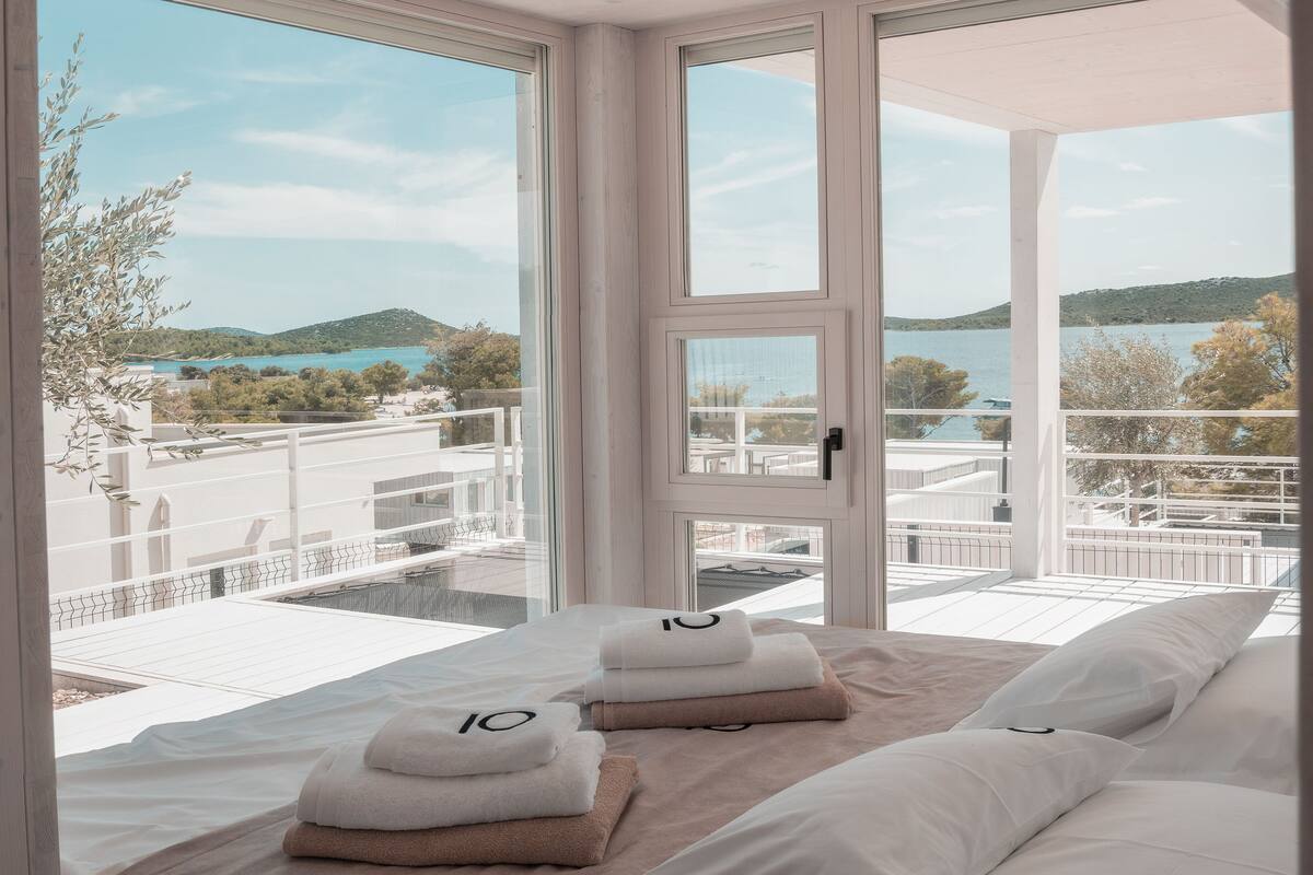 A hotel room with a large window offering a view of a coastal landscape. The room features a bed with neatly folded towels and a wooden headboard.
