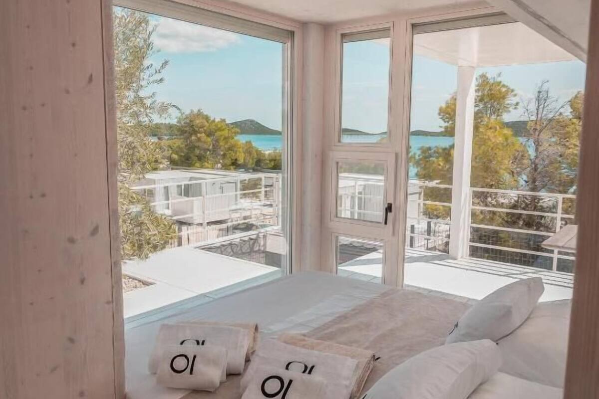 A bedroom with a large window, a bed with white linens, and several pillows with the letter "O" on them.