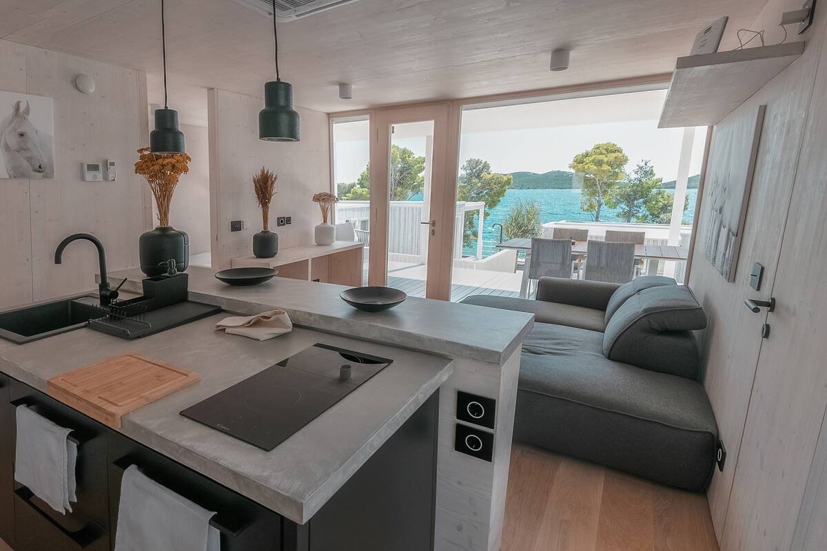 A modern kitchen with a central island, a dining area with a sofa, and a view of the sea.