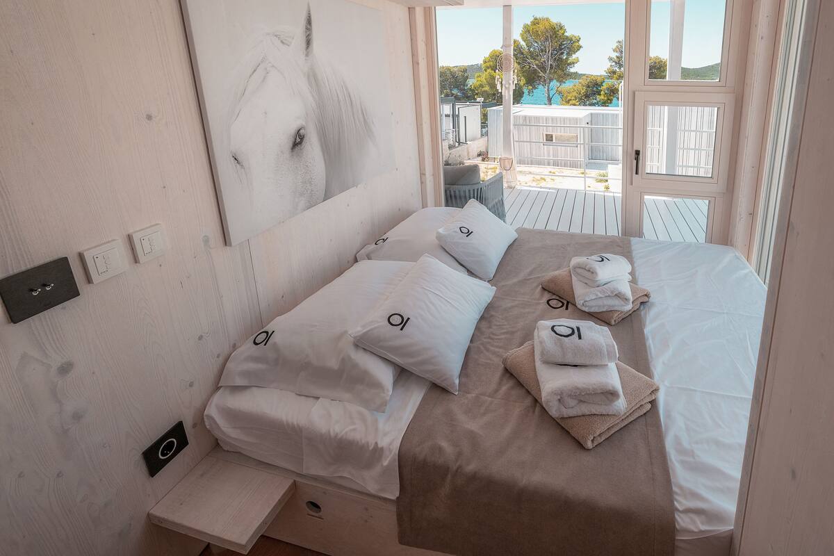 A bedroom with a large window, a bed with white and beige bedding, and a towel stack.