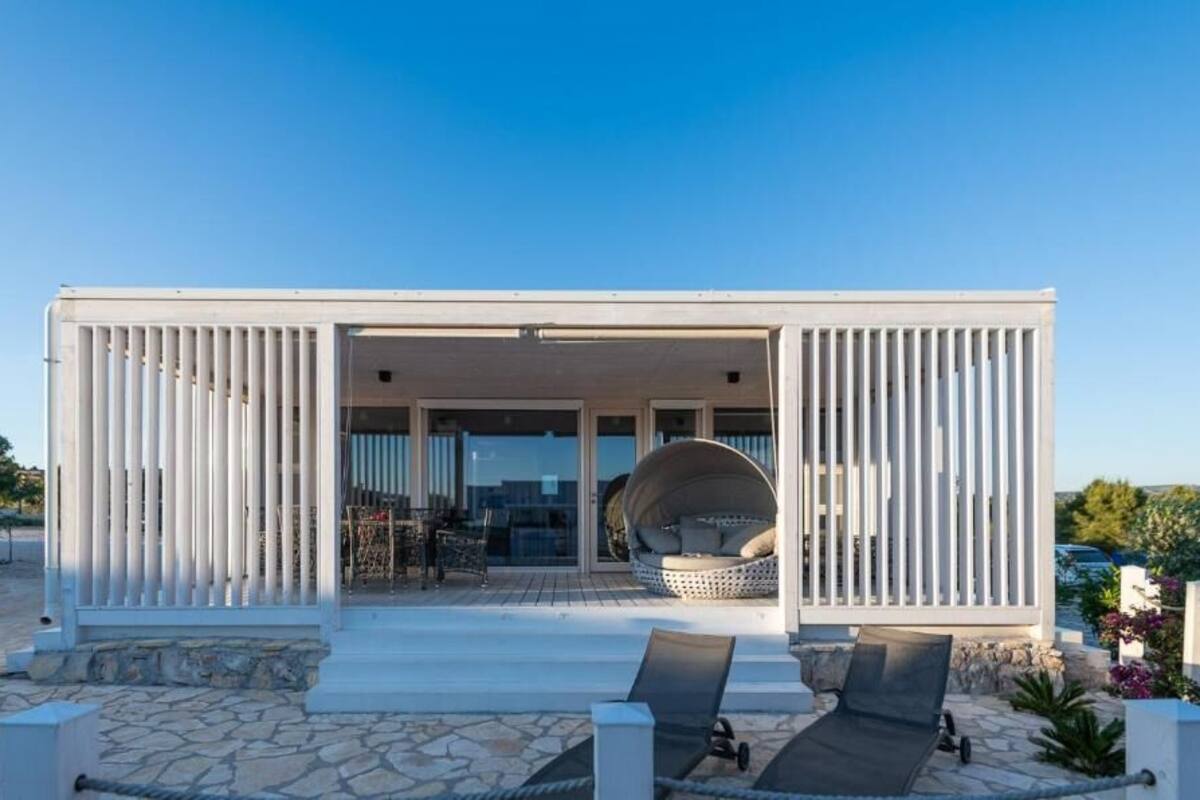 A modern, white, single-story building with a covered outdoor seating area, stone flooring, and a view of the surrounding landscape.