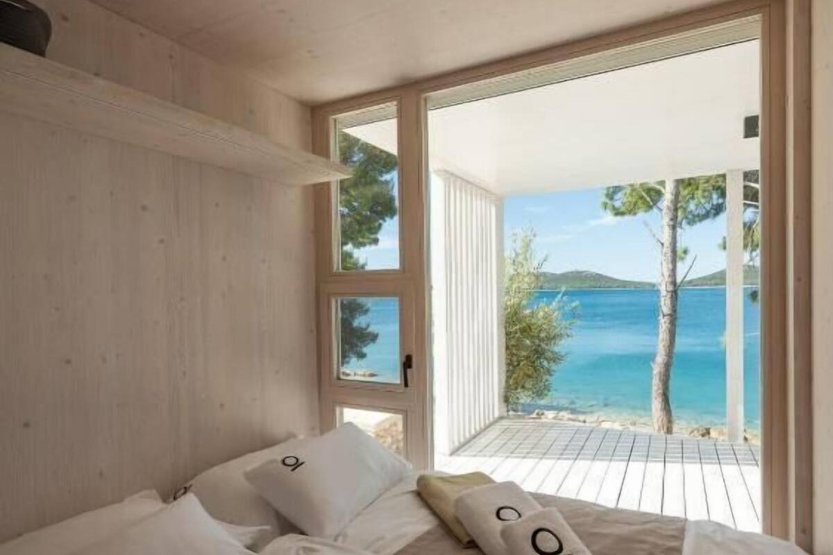 A bedroom with a large bed, white linens, and towels with a logo. It has a wooden ceiling and walls, and a view of the sea through a glass door.