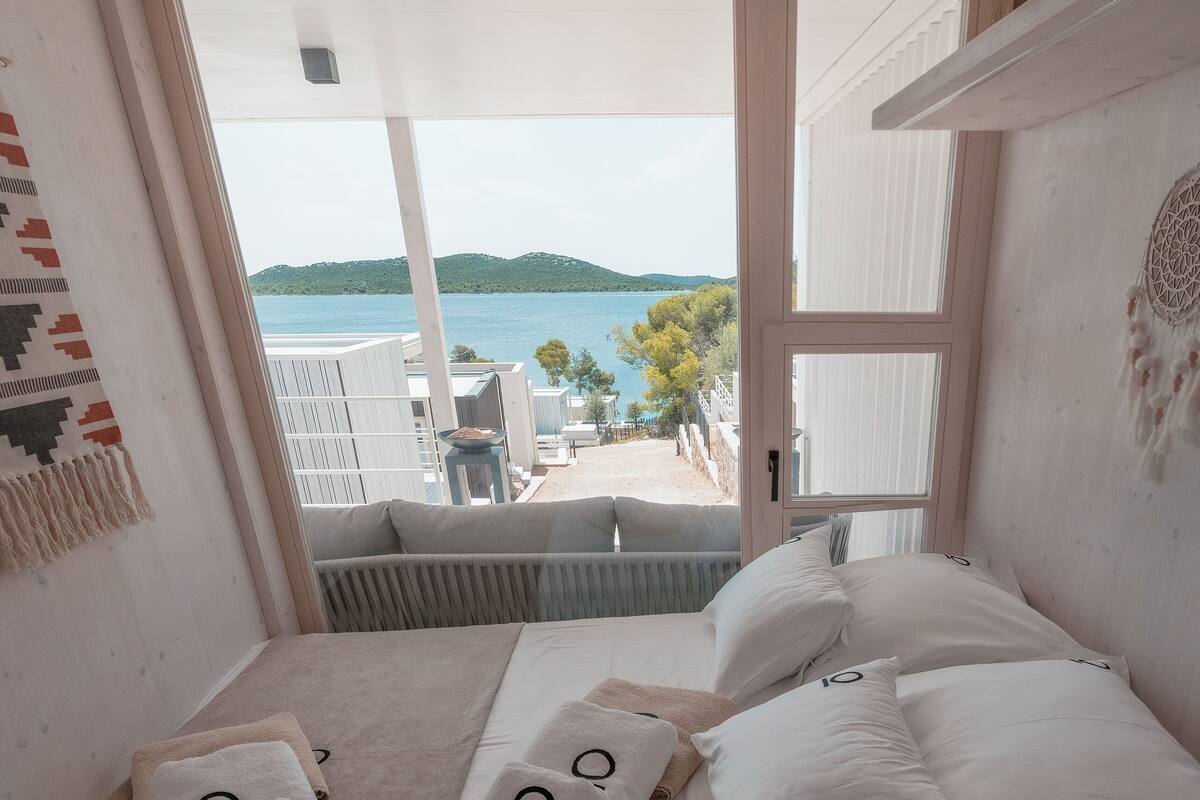 A bedroom with a large window, a bed with white bedding, and a view of the sea.