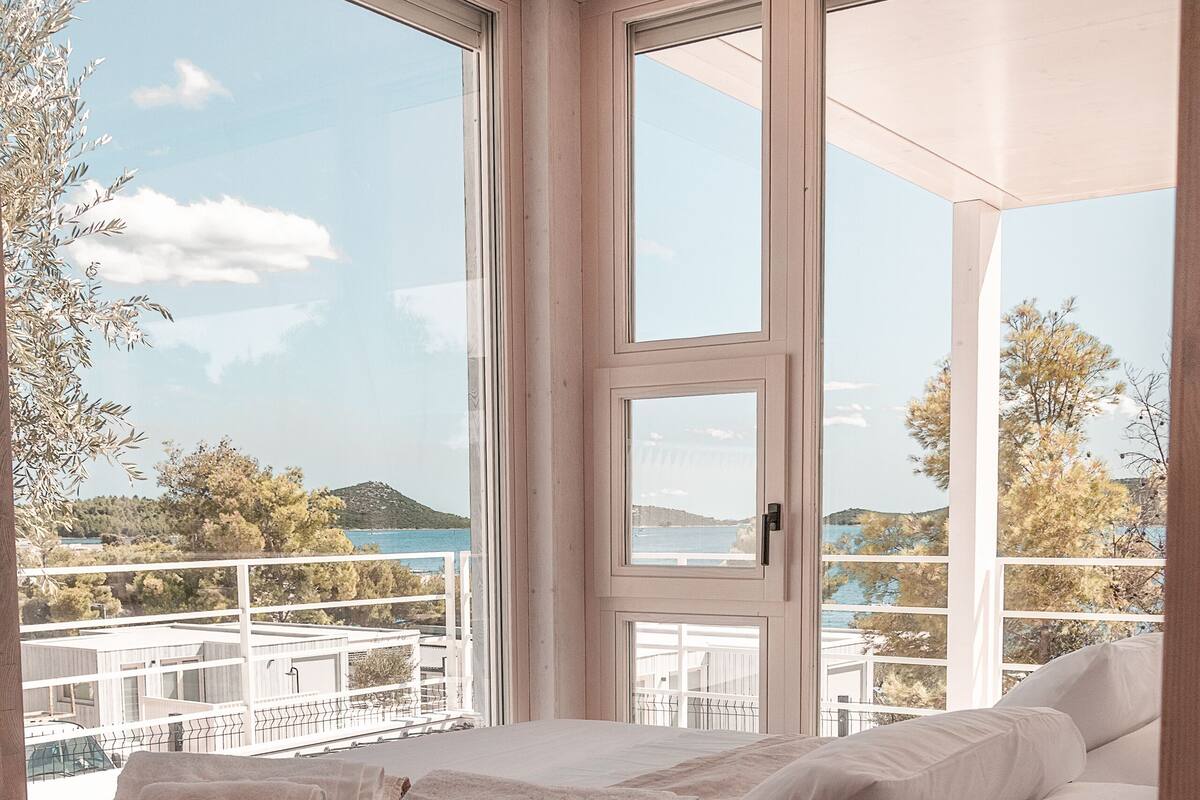 A bedroom with a large window, a bed with white linens, and a view of the sea and hills.