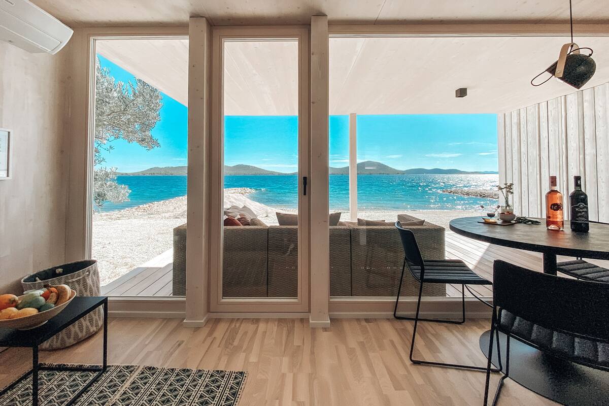 A room with a large sliding glass door leading to a beach view, a round table with chairs, and a small seating area with cushions.