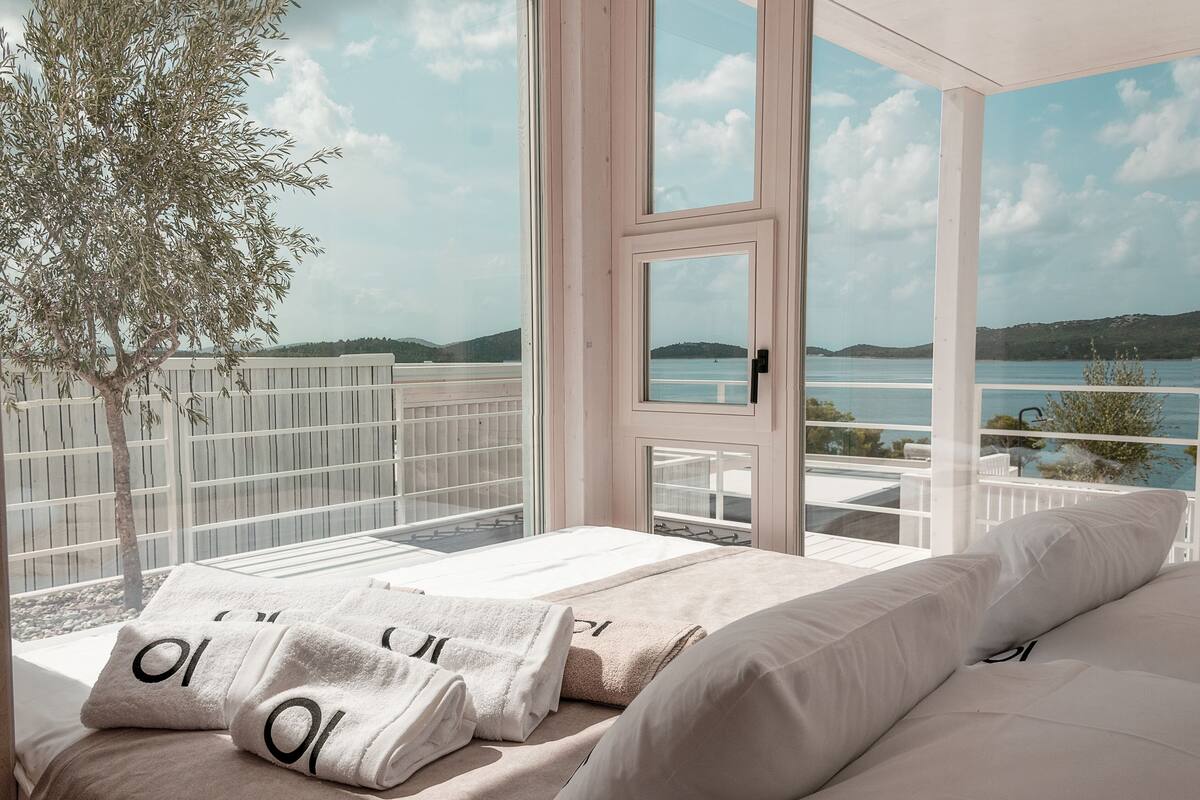 A bedroom with a large window overlooking a body of water and hills. There are two beds with white linens and towels.