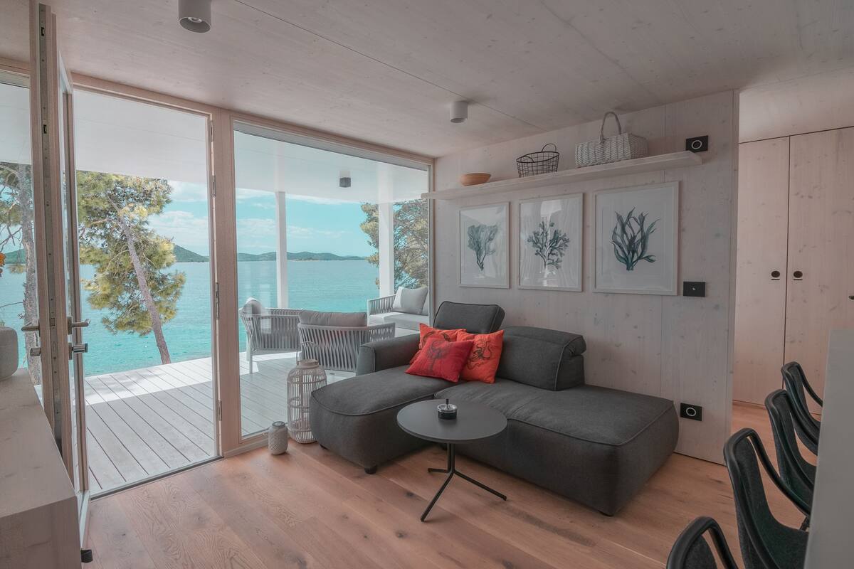 A modern living room with a grey sofa, a small round table, and a view of the sea through large windows.