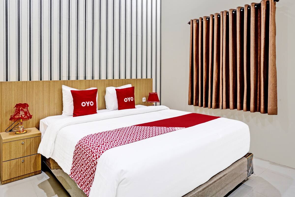 A hotel room with a wooden headboard, a bed with white and red bedding, and a red throw blanket.